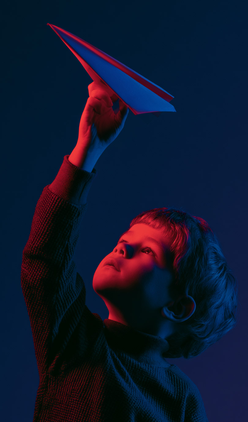 A child holding up a paper aeroplane, lit in red and blue